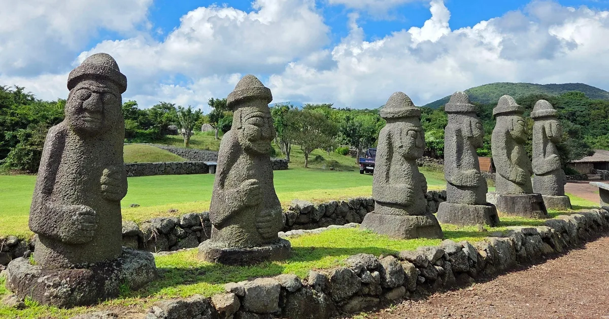 A row of carved basalt dol hareubang statues on a lawn under a blue sky, from the private taxi tour listing